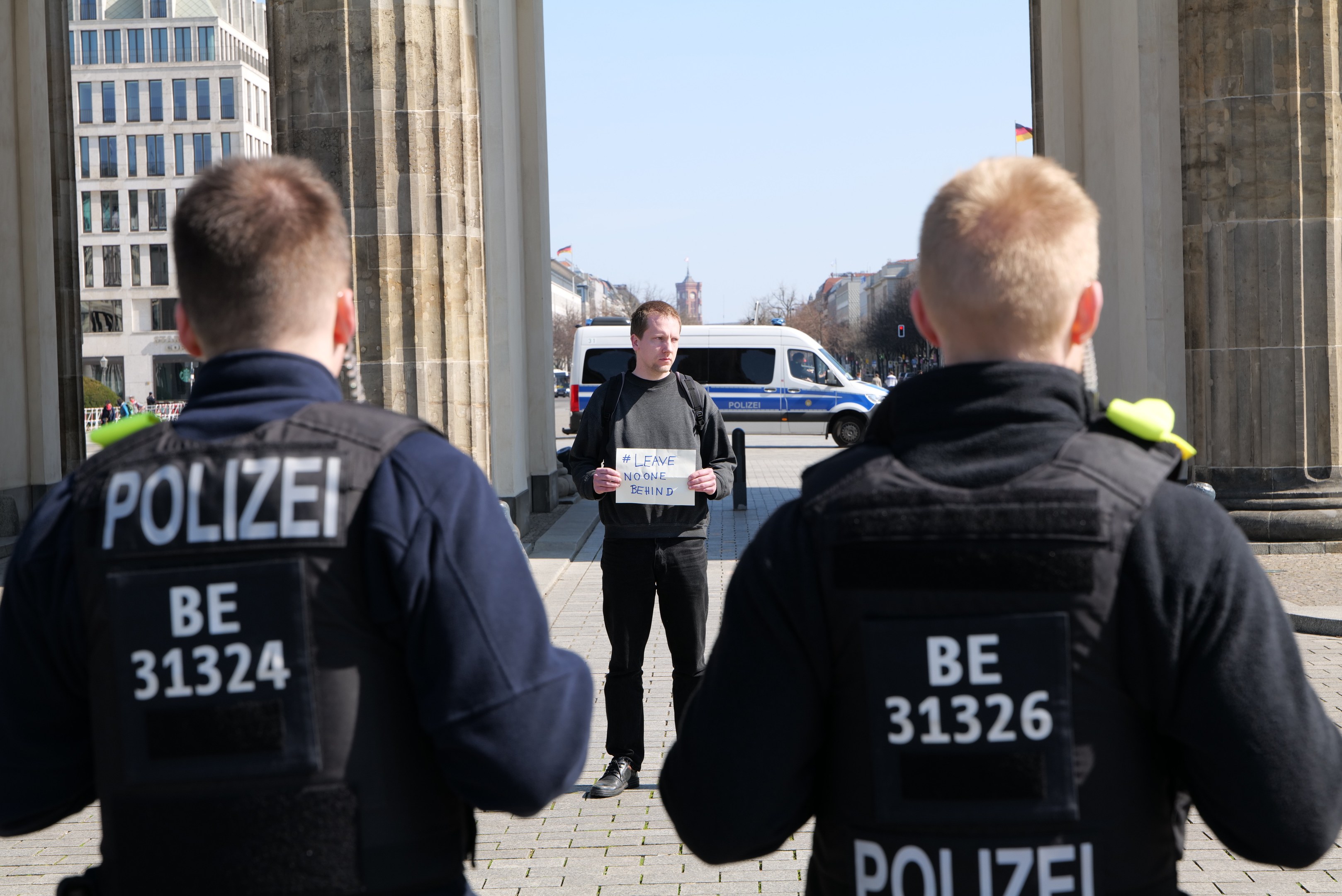 Three police officers standing in front of a building, with a man holding a paper, flanked by pillars, in a busy urban setting.