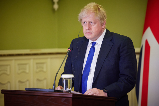 Ein Mann in Anzug und Krawatte steht an einem Rednerpult mit Mikrofon, Glas Wasser und anderen Gegenständen und spricht hinter einer Wand mit einer Flagge auf der rechten Seite.