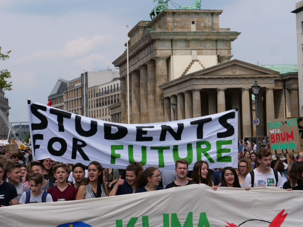 Eine Gruppe von Schülern marschiert in Berlin, hält eine bunt bemalte Tafel mit der Aufschrift "Schüler für die Zukunft", mit Gebäuden, Bäumen und Himmel im Hintergrund.