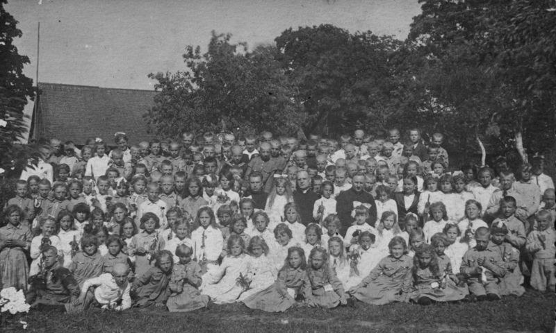 Schwarz-weißes Foto einer Gruppe von Kindern, die vor einem Haus mit Bäumen und einem klaren Himmel posieren.