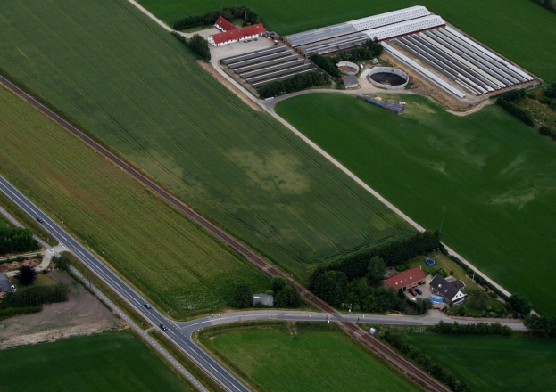 Luftaufnahme eines hydroponischen Bauernhofs in einer grünen Landschaft, mit einem zentralen Gebäude, umgeben von Bäumen, Häusern, Straßen und Fahrzeugen.