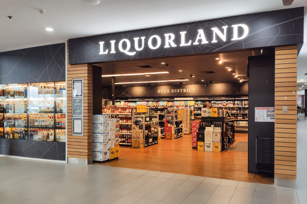 Ein Liquorland-Geschäft in einem Einkaufszentrum mit einem Schild an der Wand, Regalen voller Flaschen und Kartons, einer Glaswand mit Postern links und Lampen auf dem Dach.