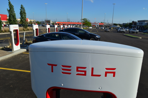 Weißes Tesla-Ladestation-Schild in einer Parkanlage mit geparkten Autos, umgeben von Gebäuden, Bäumen, Strommasten und einem klaren blauen Himmel.