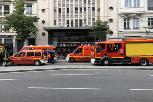 Eine Gruppe von Feuerwehrautos auf einer Straße in Paris geparkt, mit Menschen auf dem nahen Gehweg, Gebäuden, Bäumen und einem Fahrrad im Hintergrund.