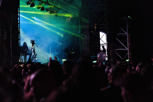 Menge bei einem Konzert mit Musikern auf der Bühne, die Instrumente spielen, unter Bühnenbeleuchtung, mit einem Banner, Metallrahmen und Mikrofonständern.