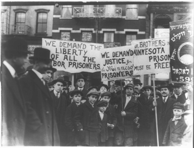 Schwarz-weiß-Fotografie einer Reihe von Menschen mit Protestschildern, auf denen "Wir fordern die Freiheit aller Gerechtigkeit für die Gefangenen" steht, vor einem Gebäude mit Fenstern und einem Geländer.