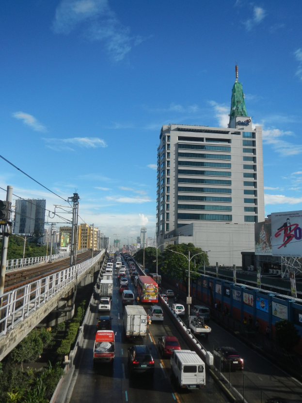 Eine belebte Stadtstraße mit hohen Gebäuden, eine Brücke auf der linken Seite, Strommasten mit Drähten, Bäume und ein bewölkter Himmel.