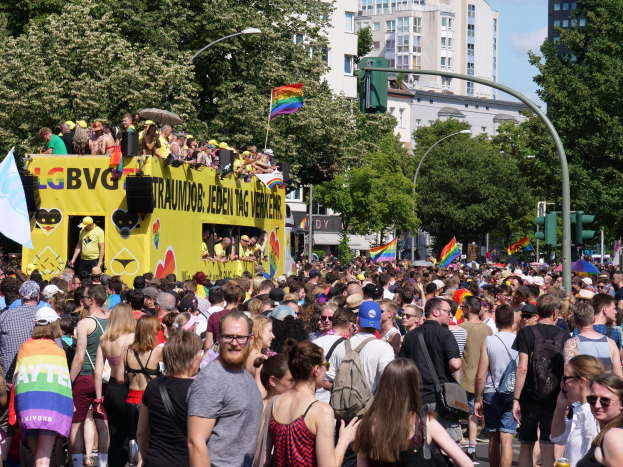 Eine große Menge marschiert auf einer Straße in Berlin, mit einem gelben Lkw im Hintergrund, einige tragen Mützen und Schutzbrillen, andere halten Fahnen, unter bewölktem Himmel mit städtischer Infrastruktur im Hintergrund.
