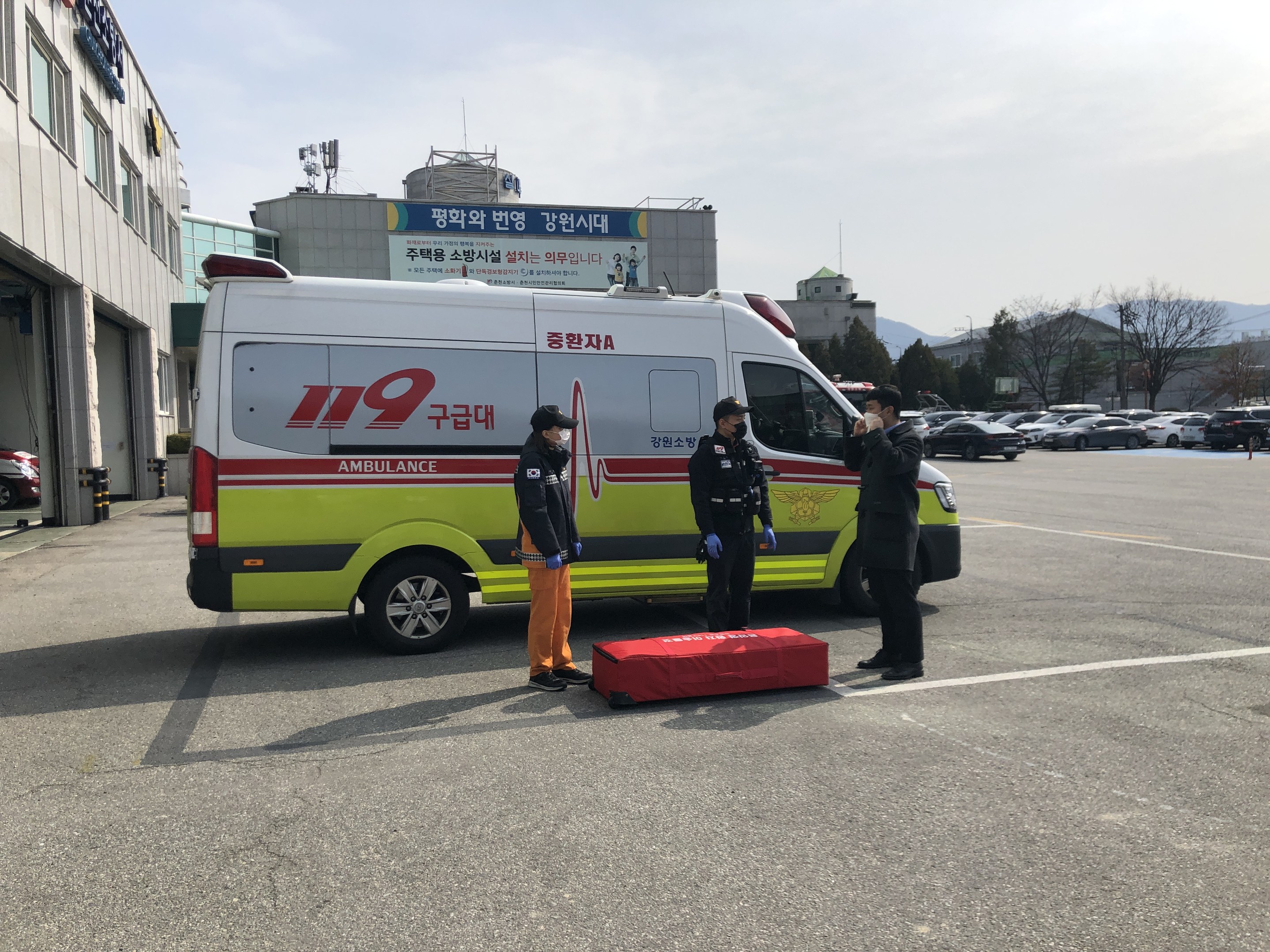 Rettungswagen auf einem Parkplatz neben einem Glasfenstergebäude mit drei Personen auf der Straße davor geparkt, andere Fahrzeuge, Bäume, Berge und Himmel im Hintergrund.