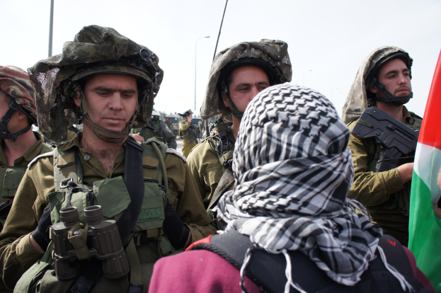 Israeli Soldaten in Uniform mit Helmen und Gewehren stehen in Formation vor einer Menge, einer hält eine Flagge, mit Laternenmasten und einem klaren Himmel im Hintergrund.