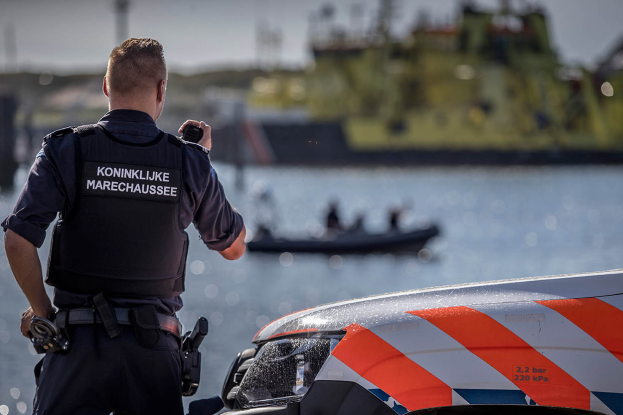 Ein Polizist in Uniform steht neben einem Polizeiwagen an einem Gewässer, hält ein Funkgerät in der Hand und sieht auf ein Boot im Hintergrund, während der Himmel darüber zu sehen ist.