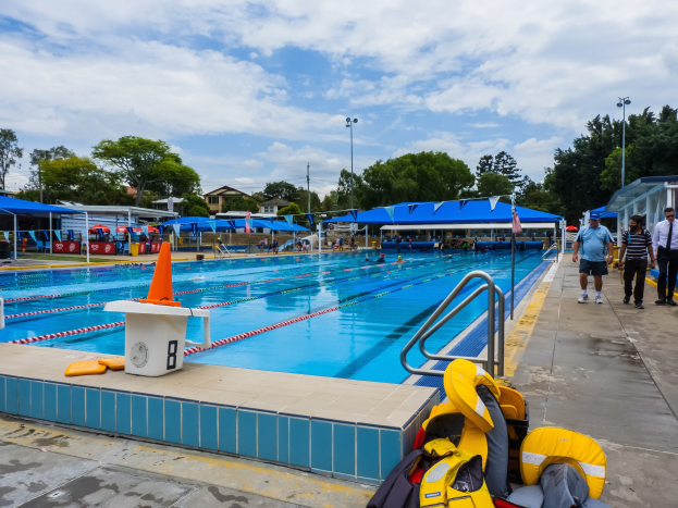 Large swimming pool surrounded by life jackets, safety cones, rods, and people, with tents, light poles, trees, buildings, and a cloudy sky in the background.