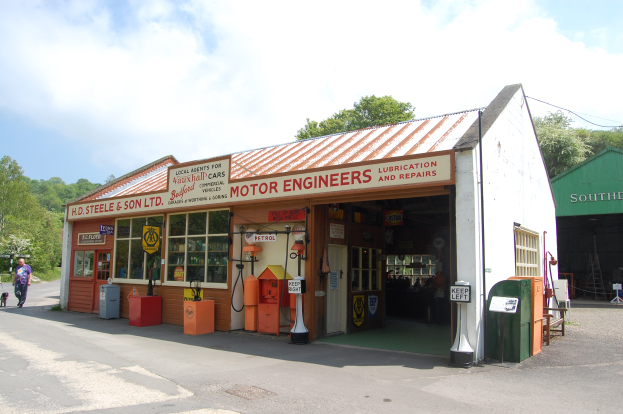 Ein Motorradwerkstatt auf der Straße mit einer Person auf der linken Seite, die eine Tasche trägt, und Bäumen mit einem klaren blauen Himmel im Hintergrund.