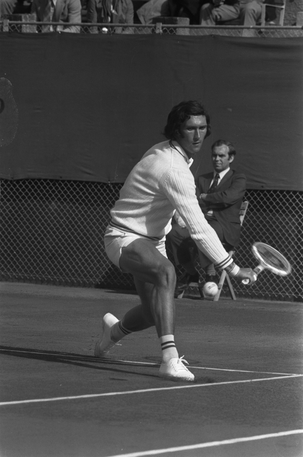 A professional tennis player in footwear swings a racquet at a ball, with mesh fencing and spectators in chairs visible in the background.