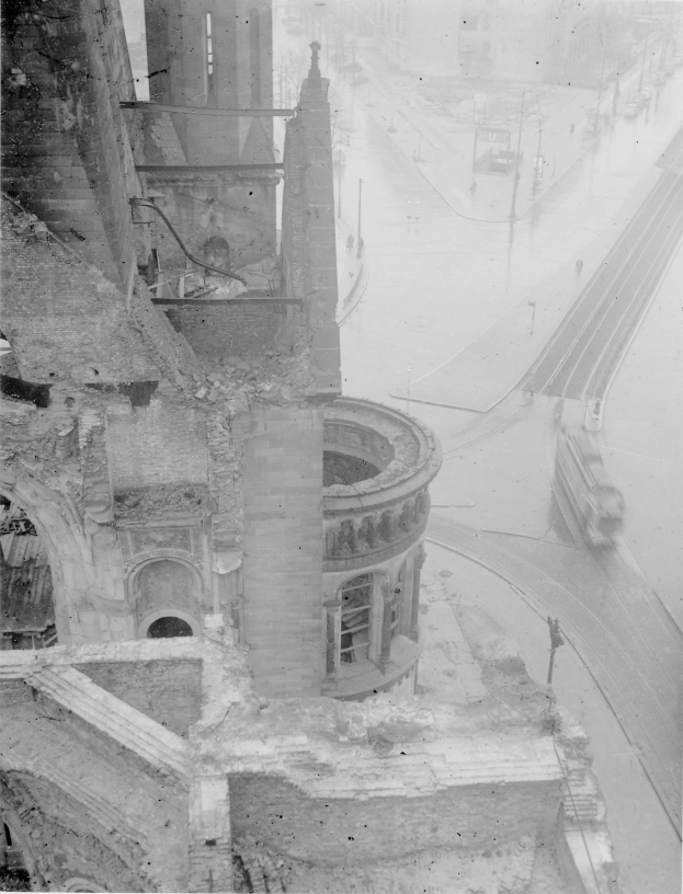 Schwarz-weiß-Foto einer Stadtstraße mit einem teilweise abgebrochenen Gebäude im Vordergrund, Fahrzeugen auf der Straße und Strommasten.