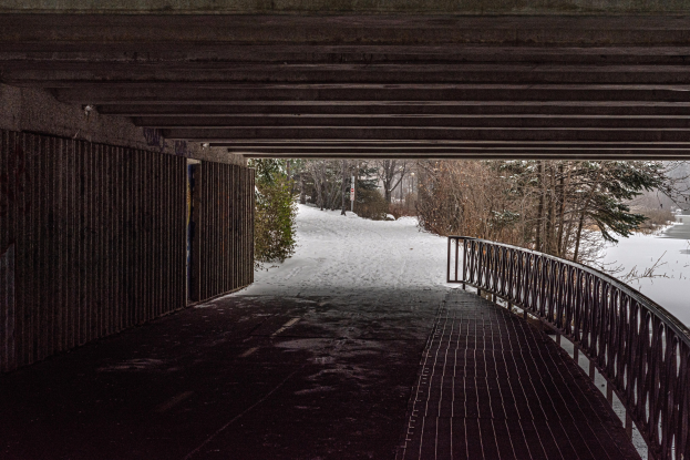 Eine schneebedeckte Straße mit einer Brücke, die Geländer und eine Wand auf der linken Seite hat, Bäume im Hintergrund und einen Wasserlauf auf der rechten Seite.