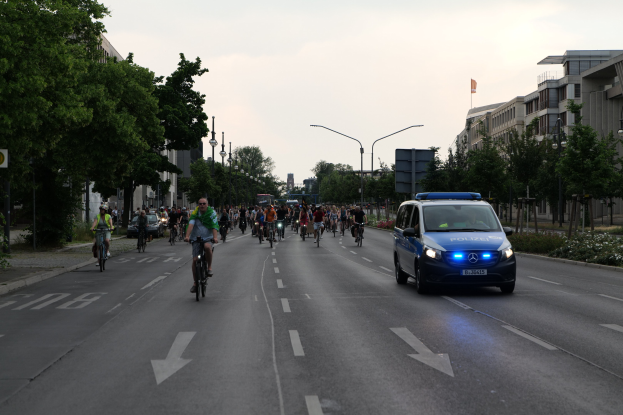 Eine Gruppe von Menschen, die auf Fahrrädern eine Straße entlangfahren, mit einem parkenden Polizeiwagen auf der Seite, flankiert von Bäumen und Gebäuden unter einem klaren blauen Himmel.