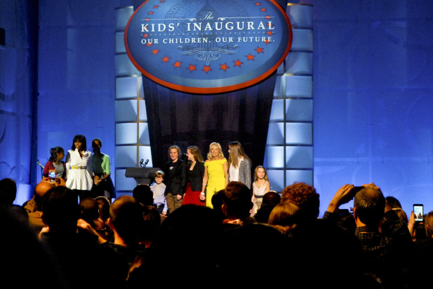 Eine Gruppe von Menschen auf einer Bühne, einige halten Mobiltelefone, mit einem Vorhang und einer Tafel mit Text im Hintergrund, die auf ein Kinderweihereignis hinweist.