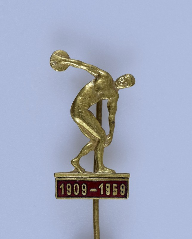 Goldmedaille mit einer Reliefabbildung eines Mannes in einem Hut, der einen Diskus wirft, mit der Inschrift "1909-1959", vor einem hellen weißen Hintergrund.