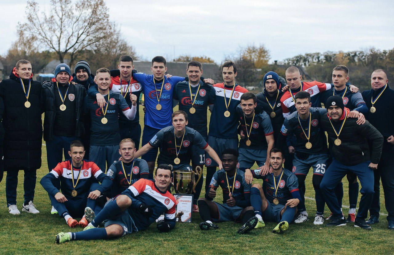 Eine Gruppe von Männern in Medaillen steht auf einem Fußballfeld mit einem Pokal, umgeben von Bäumen unter einem klaren blauen Himmel.