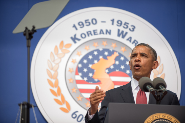 Präsident Obama in Blazer und Krawatte hält eine Rede am Korean War Memorial in Washington, D.C., vor einem Podium mit Mikrofonen, einem Banner mit Text und einem Emblem dahinter und einem Ständer links von ihm.