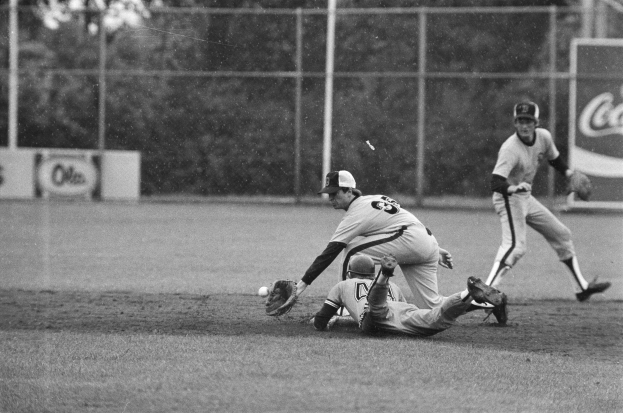 Baseballspieler rutscht beim Spiel ins Home Plate, trägt eine Kappe und Handschuhe, mit Maschendrahtzaun, Pfählen, Bäumen und Texttafeln im Hintergrund.