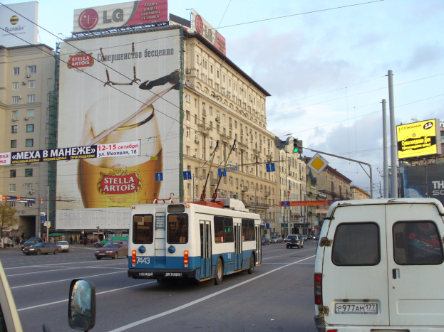 Eine belebte Stadtstraße mit mehreren Fahrzeugen, Gebäuden, Werbetafeln, Strommasten, Verkehrsampeln und einem bewölkten Himmel.