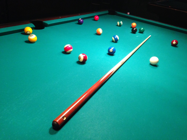 Ein grüner Poolbillardtisch mit einem Snookercue-Stock in der Mitte, umgeben von Billardkugeln in Dreiecksformation auf einem dunklen Hintergrund.