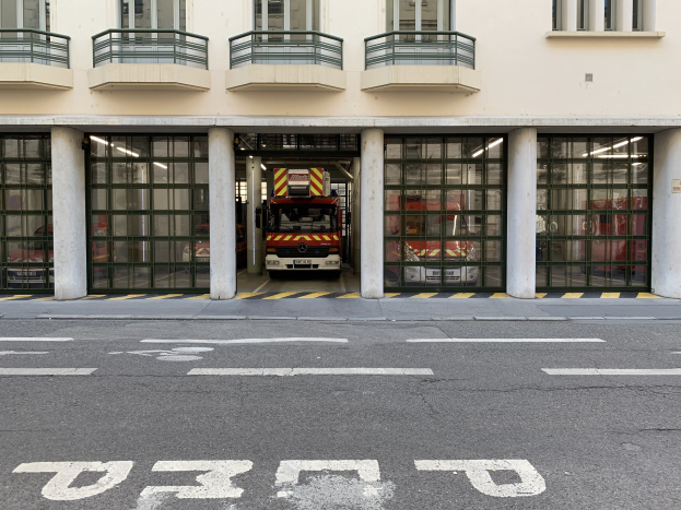 Feuerwache mit Feuerwehrauto vor dem Gebäude, mit Fenstern, Geländern, Säulen, Glastüren und einer Straße unten.