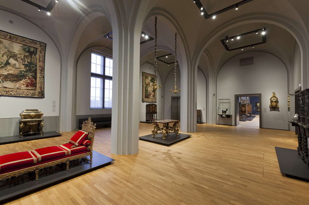Großer Raum im Kunsthistorischen Museum Wien mit Möbeln wie einem Sofa mit Kissen, Tischen mit Gegenständen, Säulen, Fenstern und Deckenleuchten sowie Gemälden an den Wänden.
