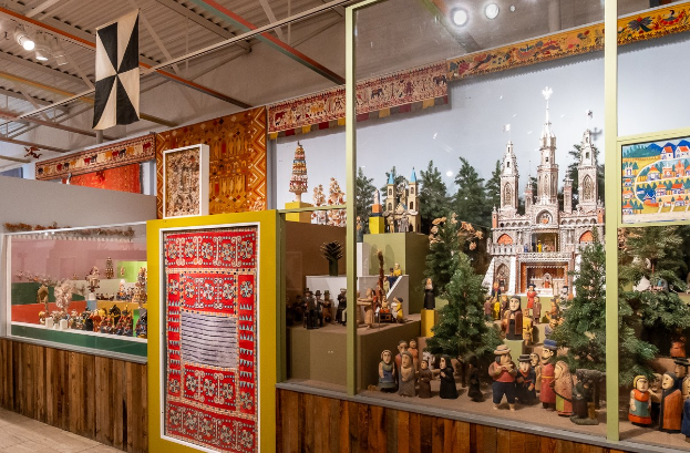 Ein unordentliches Zimmer im Museum der Russischen Föderation, das verschiedene Spielzeuge wie Puppen, Weihnachtsbäume und Gebäude zeigt, mit gerahmten Gegenständen an den Wänden und Deckenleuchten.