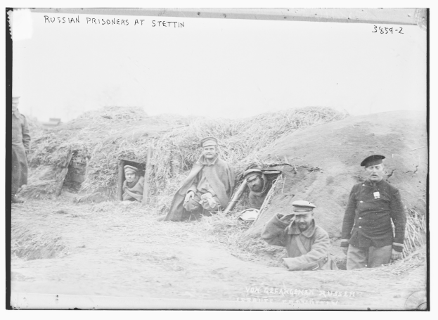 Schwarz-weißes Foto einer Gruppe russischer Gefangener, die auf einer grünen Fläche mit einem klaren Himmel im Hintergrund stehen und sitzen.