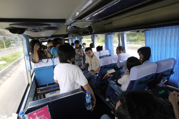 Menschen in einem Bus mit einem Zaun auf der linken Seite, einer Straßenabsperrung auf der rechten Seite, Bäumen und Pflanzen im Hintergrund, einer Person mit einer Kamera und Lautsprechern auf dem Busdach.
