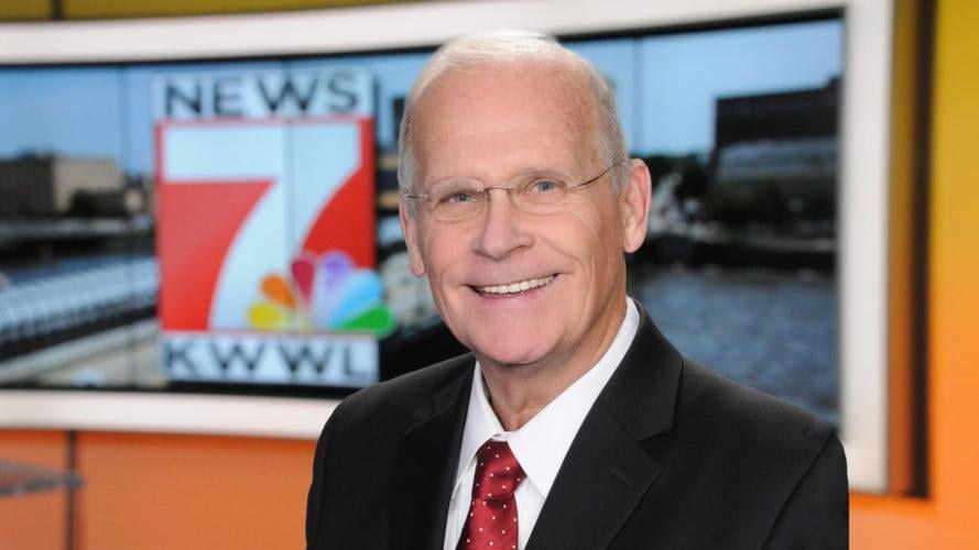 Mann im Anzug und Krawatte mit Brille vor einem Fernseher stehend, der fröhlich lächelt, wahrscheinlich ein Nachrichtensprecher.