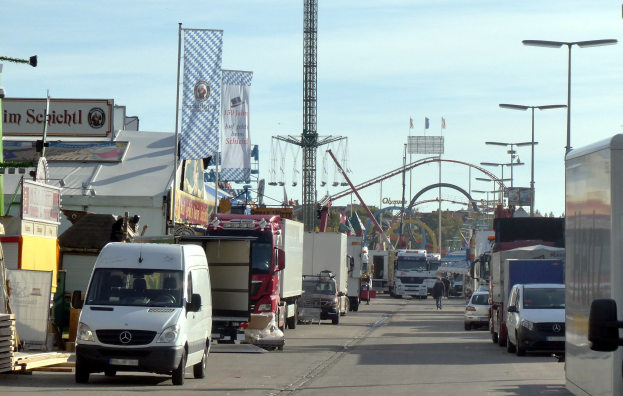 Vollgestopfte Straße mit Lastwagen, Autos, Fußgängern, Laternen, Bannern, einem Turm, einer Attraktion, Bäumen und einem bewölkten Himmel.
