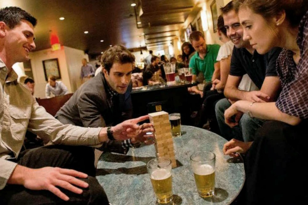 Eine Gruppe von Menschen sitzt um einen Tisch und spielt ein Biertrinkspiel, mit Gläsern Bier auf dem Tisch und Fotorahmen an der Wand im Hintergrund unter warmem Deckenlicht.