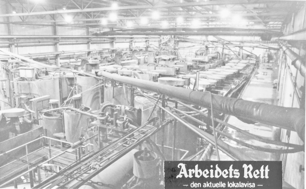 Black and white photo of an industrial factory interior with machinery, pipes, rods, and ceiling lights, featuring text at the bottom reading "Arbeibets Rett - den Aktuelle Lokalavisa".
