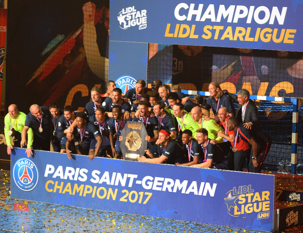 Spieler von Paris Saint-Germain feiern mit dem Ligue-1-Pokal, tragen Medaillen, stehen vor einer Tafel mit Text und Logos, mit einem Netz auf der rechten Seite und einem Bildschirm im Hintergrund.