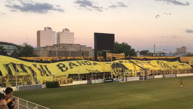 Fußballfeld mit einer Gruppe von Menschen, einem Torpfosten, einer 'Boca Juniors gegen Real Sociedad'-Plakette, Pflanzen, einem Anzeigebildschirm, Pfählen, Bäumen, Gebäuden und einem Metallzaun unter einem bewölkten Himmel.