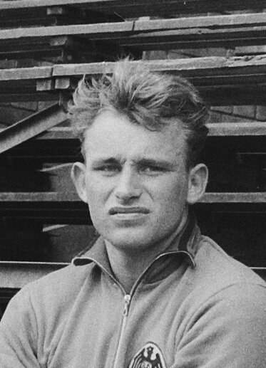 Schwarzes und weißes Foto eines Mannes in einer Jacke, der vor gestapelten Holzplanken steht, der legendäre deutsche Fußballspieler.