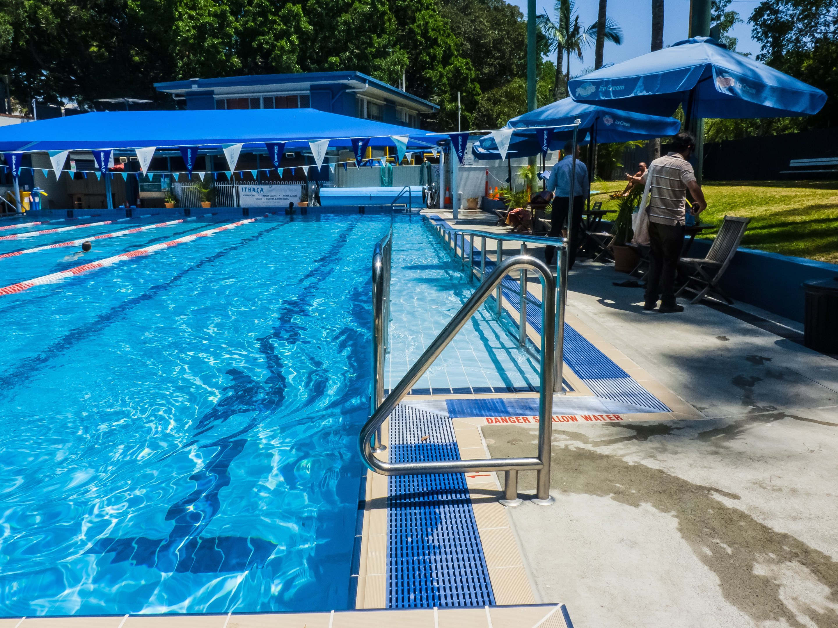 Großer Freibadpool mit schwimmenden Menschen, umgeben von Geländern, Sonnenschirmen, Stühlen, Tischen, Pflanzen, Gras, Bäumen und einem Gebäude unter einem klaren blauen Himmel.