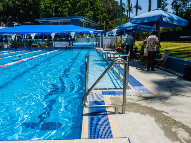 Großer Freibadpool mit schwimmenden Menschen, umgeben von Geländern, Sonnenschirmen, Stühlen, Tischen, Pflanzen, Gras, Bäumen und einem Gebäude unter einem klaren blauen Himmel.
