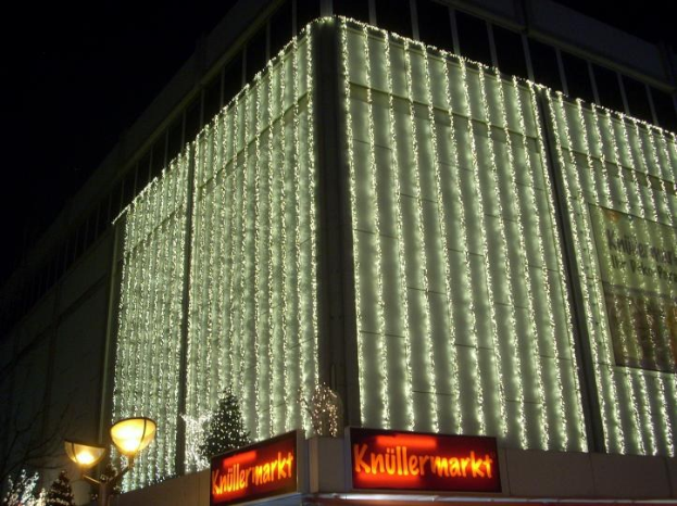 Ein Gebäude in Berlin, Deutschland, geschmückt mit festlichen Weihnachtslichtern, umgeben von Bäumen und Laternenpfählen, mit Schildern mit Text und einem sichtbaren Himmel im Hintergrund.
