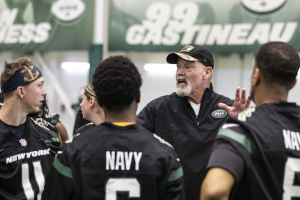 Gruppe von Männern in einem Stadion, mit Jets Head Coach Mike McCarthy in einer Mütze, der zu seinem Team spricht, umgeben von Bannern und Lichtern.