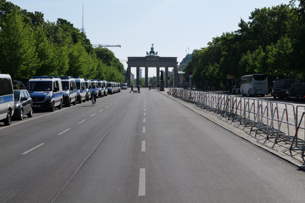 Lange Reihe von Polizeiwagen, die an einer Straße vor dem Brandenburger Tor geparkt sind, mit Radfahrern und Fußgängern, Absperrungen, Bäumen und einem Bogen mit Statuen im Hintergrund.