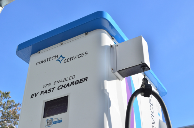 Elektrische Fahrzeug-Ladestation mit einem blauen und weißen "V2G Fast Charger"-Schild, umgeben von Bäumen und einem Gebäude unter einem klaren blauen Himmel.