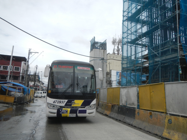 Ein Bus fährt auf einer Stadtstraße mit hohen Gebäuden, Baustelle auf der rechten Seite, anderen Fahrzeugen auf der linken Seite, Strommasten, Drähten und Himmel im Hintergrund.