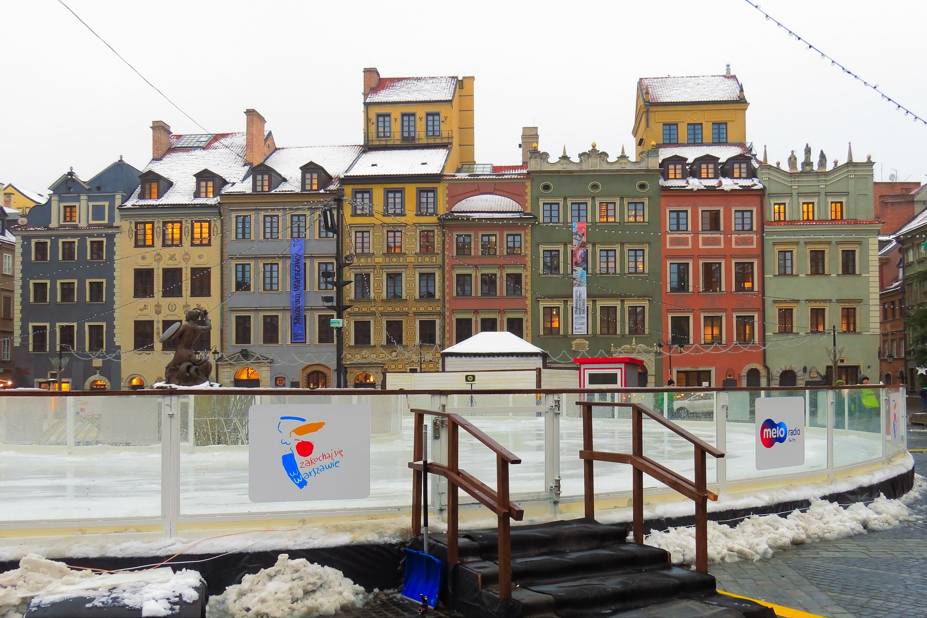 Eislaufbahn in einem Stadtplatz umgeben von Geb├Ąuden, Treppen, Gel├Ąndern, Informationsschildern, schneebedecktem Boden, Laternen, B├Ąumen und einem Himmel als Hintergrund.
