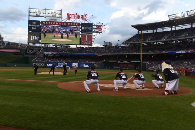 Eine Gruppe Baseball-Spieler konkurriert in einem Spiel im Nationals Park, mit Zuschauern auf den Tribünen und einem großen Bildschirm im Hintergrund bei teilweise bewölktem Himmel.