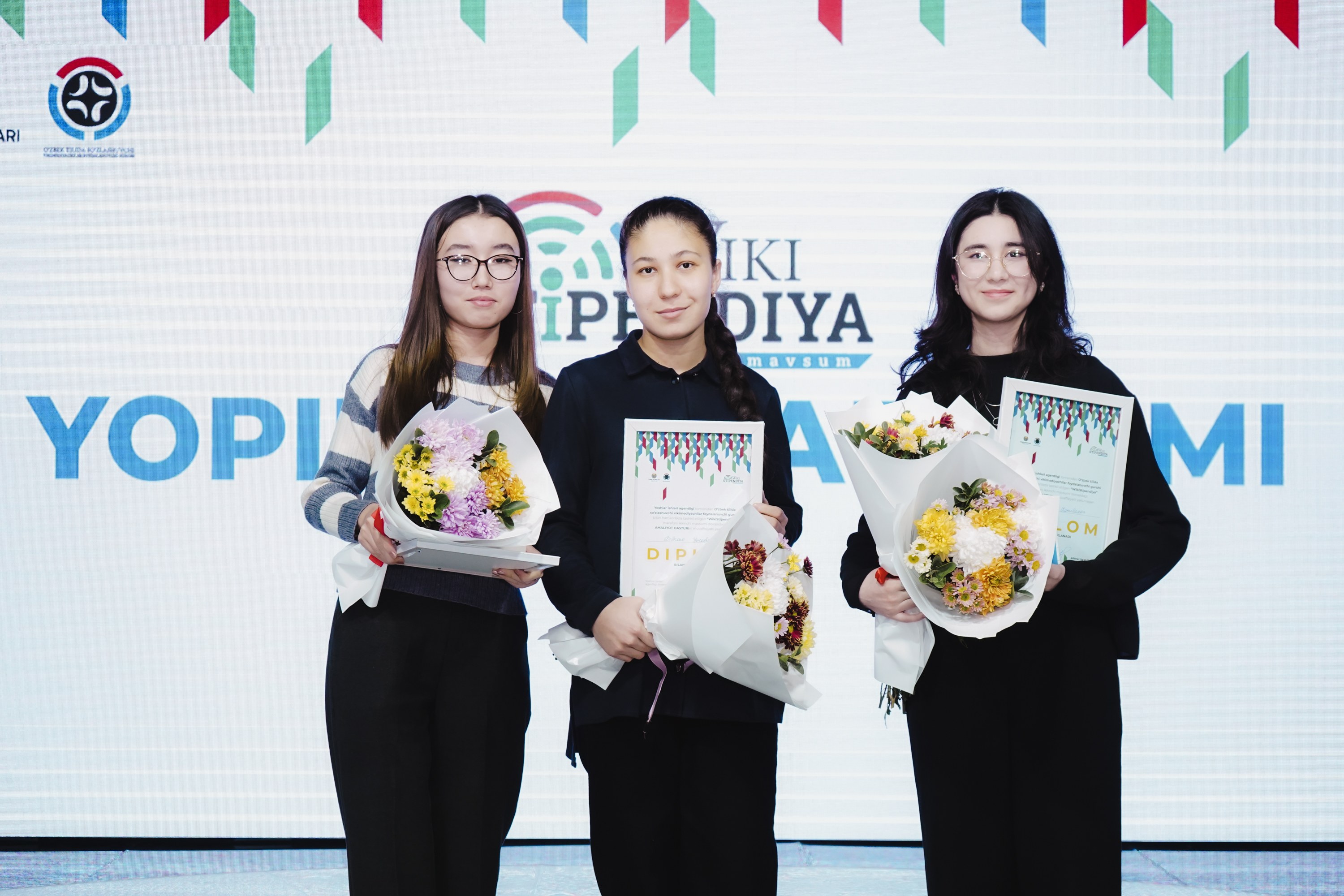 Drei Frauen mit Blumensträußen und Urkunden auf einer Bühne mit einem "Yopi Diya Awards 2019"-Plakat im Hintergrund.